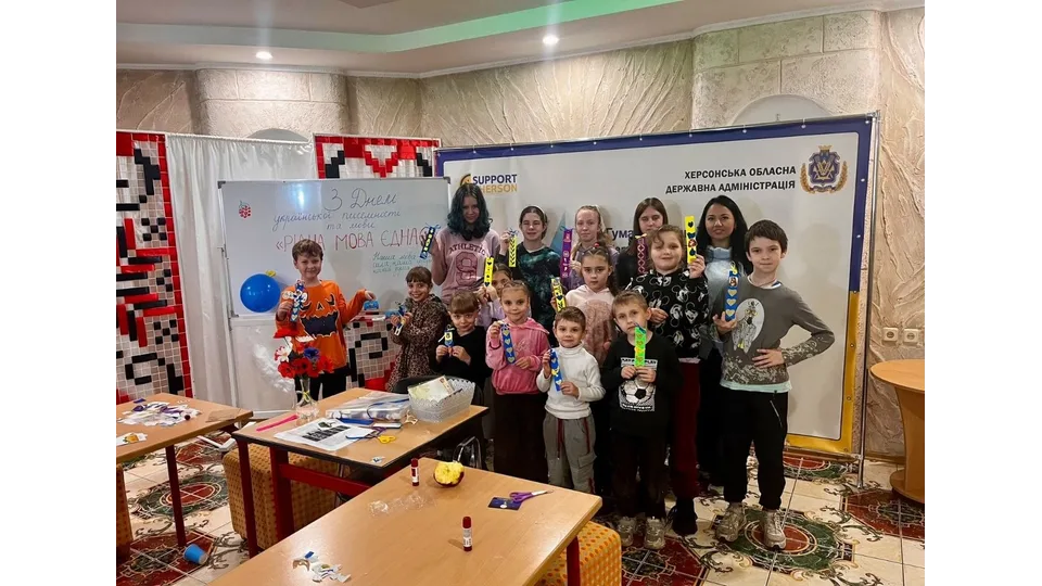Спільне фото учасників дитячого творчого заходу «Рідна мова єднає» до Дня української писемності та мови в Одеському координаційному центрі «Вільні разом»: у залі з візерунчастим кахельним покриттям підлоги стоїть велика група дітей-переселенців з Херсонщини разом із двома дорослими жінками-організаторами, які посміхаються та тримають у руках виготовлені під час майстер-класу паперові синьо-жовті та різнокольорові закладки для підручників; ліворуч стоїть велика магнітна біла дошка із написом від руки «З Днем української писемності та мови. «РІДНА МОВА ЄДНАЄ», паперовими синьо-жовтими серцями та прикріпленою синьою повітряною кулькою, а на задньому плані праворуч встановлено великий темно-синій банер «ХЕРСОНСЬКА ОБЛАСНА ДЕРЖАВНА АДМІНІСТРАЦІЯ» з гербом області та логотипом благодійного фонду «SUPPORT KHERSON»; на передньому плані перед дітьми розставлено низькі дитячі столики з матеріалами для творчості, ножицями, клеєм та папером, а з лівого боку приміщення завішане білою ширмою з традиційним червоно-чорним українським вишитим орнаментом.