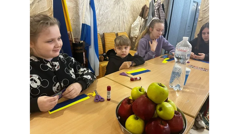 Діти-переселенці з Херсонщини під час творчого майстер-класу в Одеському координаційному центрі «Вільні разом»: за довгим дерев'яним столом сидить група дітей, які займаються виготовленням паперових поробок та закладок для книг; безпосередньо на передньому плані ліворуч усміхається дівчинка в чорній кофті з малюнками Міккі Мауса, перед якою на столі лежать ножиці, клей-олівець та синьо-жовта паперова основа; по центру столу стоїть велика металева миска, наповнена стиглими червоними та зеленими яблуками, а поруч — велика пластикова пляшка мінеральної води «Моршинська»; за столом далі сидять маленький хлопчик у чорному худі, дівчина у бузковій кофті з резинкою-бантом на волоссі та ще одна дівчинка праворуч, яка розглядає кольорові картки; на задньому плані ліворуч у кутку кімнати встановлено на стійках державний прапор України та прапор Херсонської області, а вздовж стіни стоять м'які стільці з помаранчевою оббивкою та висить верхній одяг.