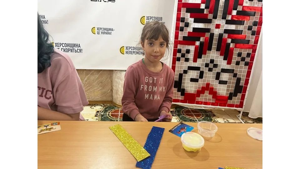 Маленька учасниця творчого заходу «Рідна мова єднає» під час майстер-класу в Одеському координаційному центрі «Вільні разом»: по центру за дерев'яним столом сидить дівчинка з челкою та зібраним волоссям у рожевому світшоті з білим текстовим принтом «GOT IT FROM MY MAMA», яка посміхається та дивиться в камеру, тримаючи в руках матеріали для поробки; на столі перед нею викладено блискучі заготовки для паперової синьо-жовтої закладки у формі галочки, кольорову ігрову картку та круглу прозору баночку з пластиліном або масою для ліплення; на задньому плані ліворуч стоїть білий інформаційний стенд із патріотичними гаслами «ХЕРСОНЩИНА НЕПЕРЕМОЖНА!», «ХЕРСОНЩИНА — ЦЕ УКРАЇНА!» та «ХЕРСОНЩИНА СКОРИТЬСЯ!», а праворуч облаштовано велике декоративне панно на стійці з традиційним українським геометричним вишитим орнаментом у червоно-чорних та білих кольорах.